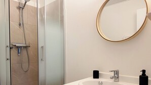 Een badkamer met een douche met glazen wand, een witte wastafel met een ronde spiegel, en twee zwarte zeepdispensers.