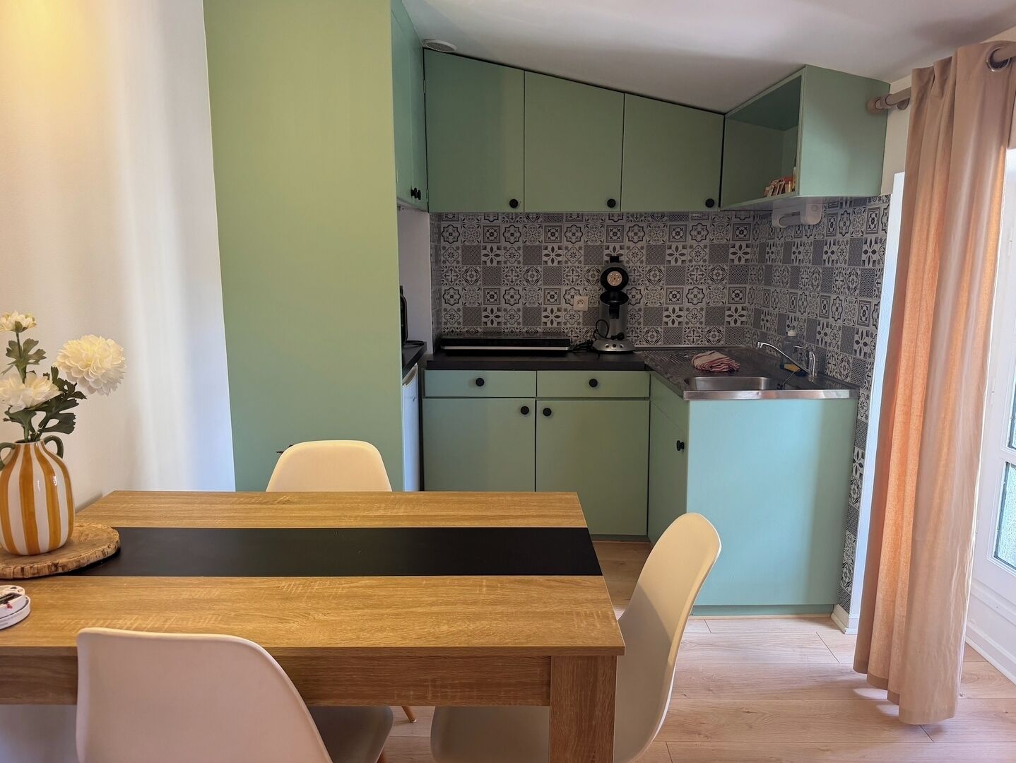 Un comedor con mesa y sillas de madera, una cocina con armarios verde claro y salpicadero decorado, y un ventanal con cortinas.