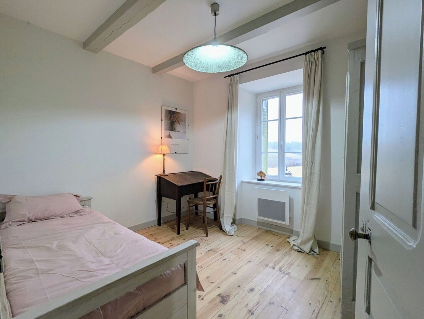 Une chambre à coucher comprenant un lit, un bureau, une chaise, une fenêtre avec des rideaux et un luminaire suspendu.