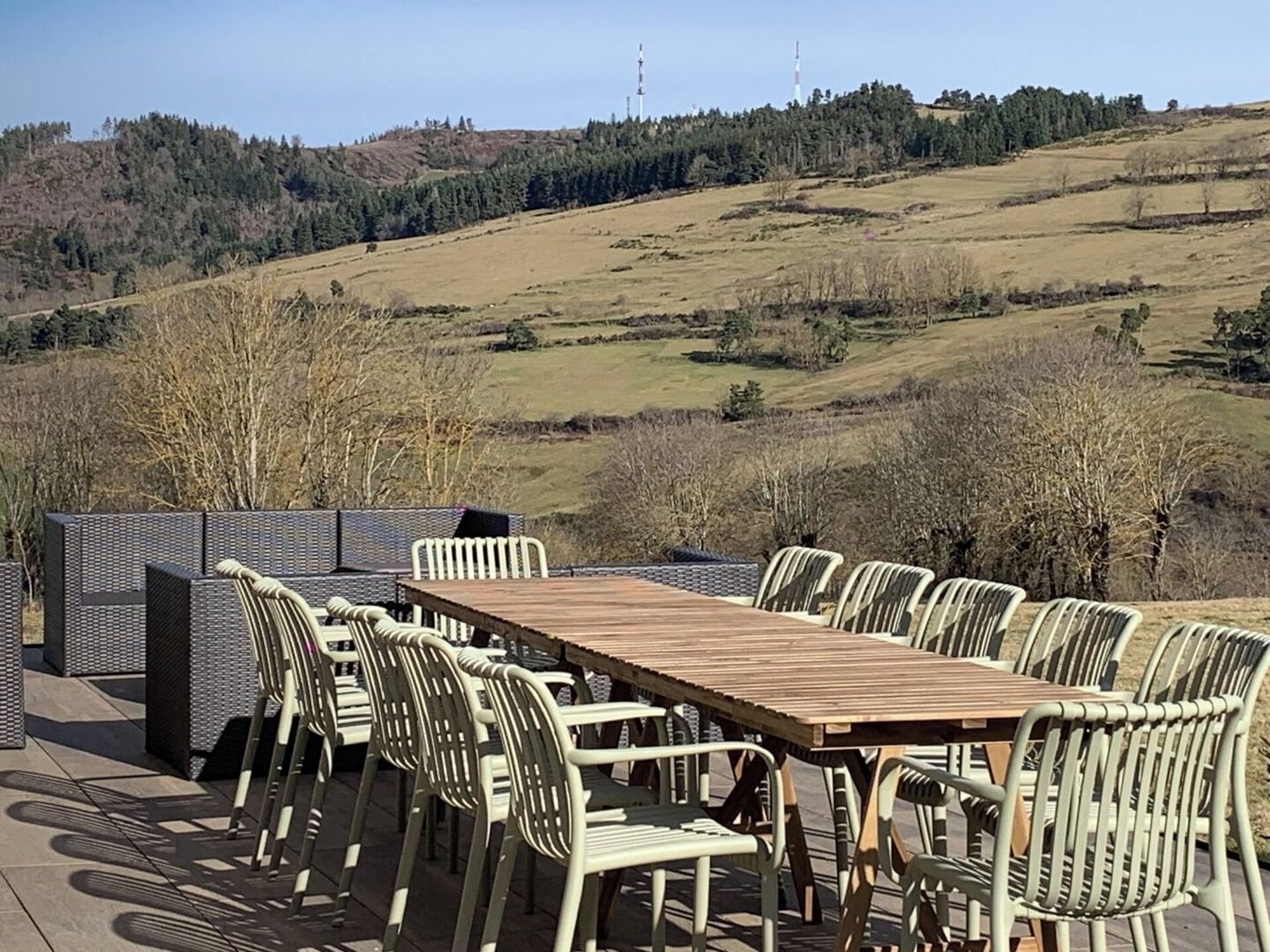 Un espace de restauration en plein air avec une longue table en bois et plusieurs chaises, le tout agrémenté d’un paysage de collines douces et de quelques arbres clairsemés.