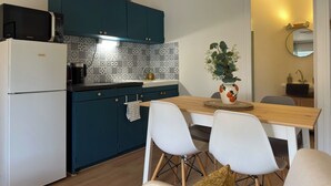 Une cuisine compacte avec des meubles de couleur turquoise, un four à micro-ondes Samsung, un réfrigérateur blanc et un coin repas comprenant une table en bois et des chaises blanches.