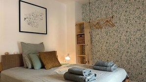 Un lit bien fait, avec une couverture et des oreillers gris, une table de chevet en bois surmontée d’une lampe, et un mur recouvert d’un papier peint à motifs floraux.