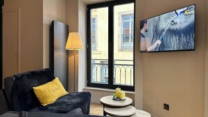 A modern hotel room with a dark blue sofa, a yellow pillow, a floor lamp, a small round table with candles, and a TV mounted on the wall.