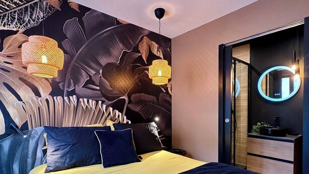 A modern bedroom with a large bed, dark blue bedding, and a wall mural of leaves. There are two hanging lamps and a round mirror on the wall.