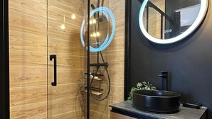 A modern bathroom with a glass-enclosed shower, a round mirror with a light, and a wooden vanity with a black sink.