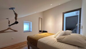 A modern bedroom with a wooden floor, a bed with white and beige bedding, a window, a mirror, and a cat tree. - Le rocher des Costes (Le Puy)