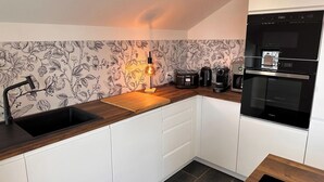 A modern kitchen with white cabinets, a wooden countertop, a black sink, a built-in oven, a coffee maker, and a vintage-style lamp. - Le rocher des Costes (Le Puy)