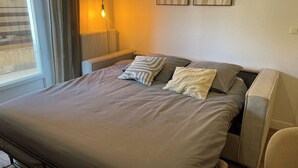 Ein ordentlich bezogenes Bett mit grauer Tagesdecke und zwei Kissen, ein Nachttisch mit Lampe sowie zwei an der Wand hängende abstrakte Gemälde.