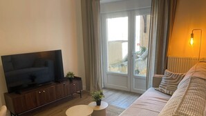 Ein Wohnzimmer mit Flachbildfernseher auf einer Holzkassette, einer Couch mit Kissen, einem kleinen runden Tisch und einer Topfpflanze.