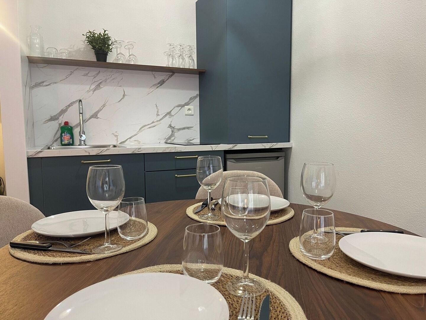 Een eetgedeelte met een tafel gedekt voor vier personen, inclusief borden, glazen en bestek. Achtergrond toont een keuken met donkere kastjes en een marmeren achterwand.