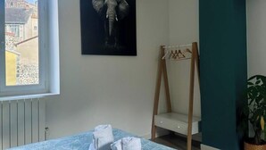 Une chambre d’hôtel avec un lit, une fenêtre donnant sur des immeubles, une œuvre d’art représentant un éléphant accrochée au mur, une chaise en bois et une plante en pot.