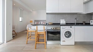 A modern kitchen with white cabinets, a washing machine, and a dining area with two chairs. - Step To The Sea 5, by Pausa Holiday Rentals (Funchal)