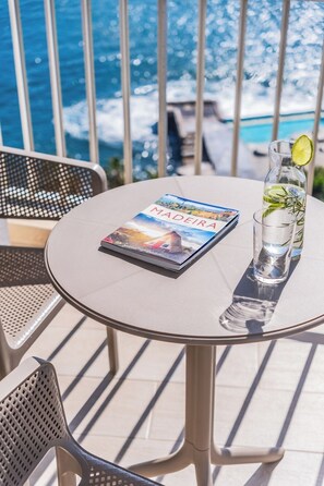 A round table with a magazine titled 'MADEIRA', a glass of water with a lime slice, and a bottle with a lime slice on a balcony overlooking the ocean. - Step To The Sea 5, by Pausa Holiday Rentals (Funchal)