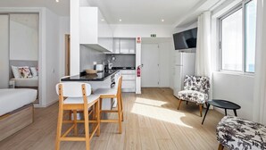 A modern, well-lit room with a kitchen area, a dining table with chairs, a sofa, and a TV mounted on the wall. - Step To The Sea 5, by Pausa Holiday Rentals (Funchal)