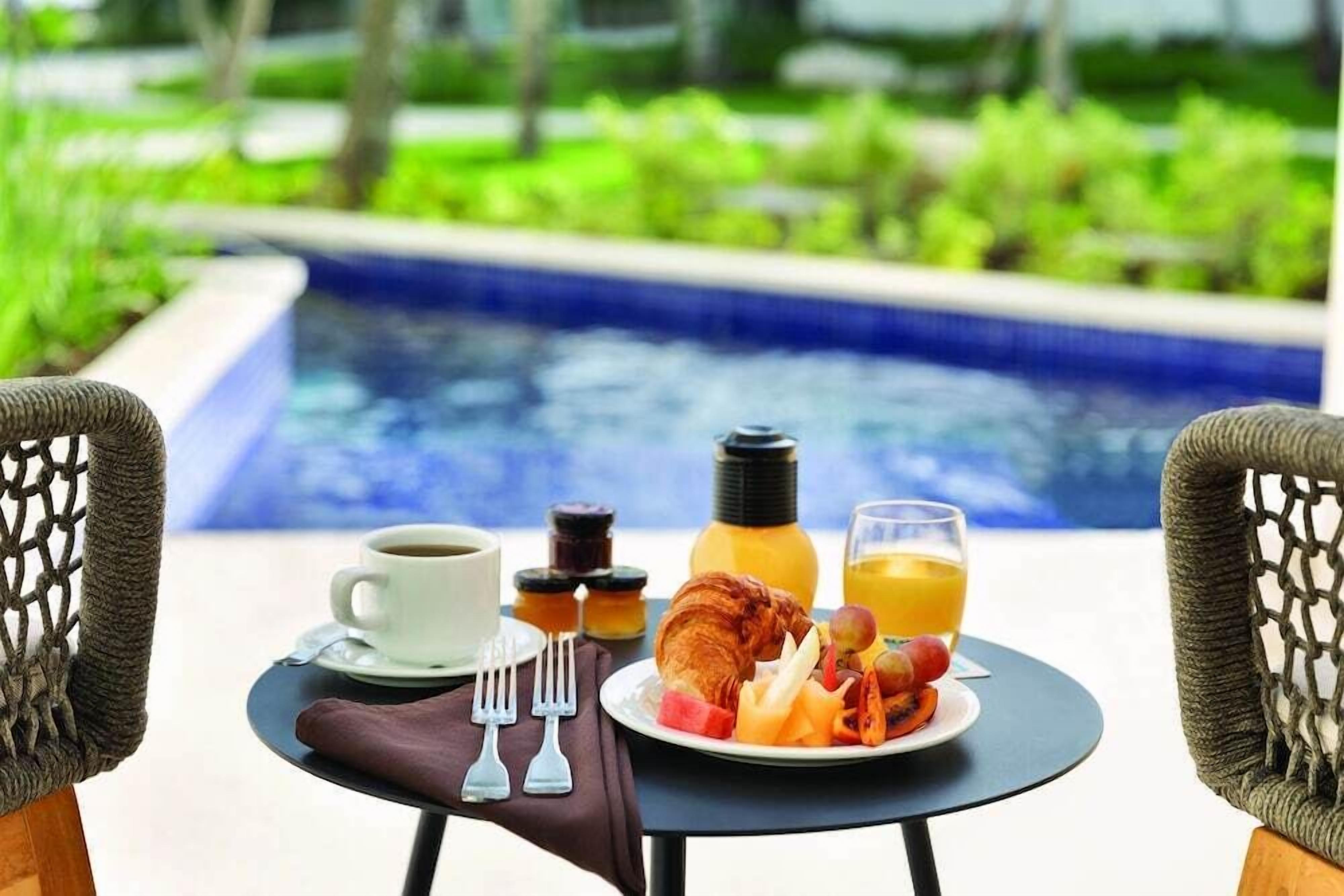 Desayuno con croissant, fruta y zumo de naranja sobre una mesa junto a una piscina.