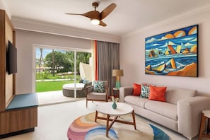A modern living room with a sofa, armchair, and coffee table. There is a painting on the wall, a ceiling fan, and a TV mounted on the wall. - Dreams La Romana All Inclusive Resort & Spa (San Rafael del Yuma)
