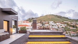 A modern rooftop terrace with a wooden deck, a built-in seating area, and a view of a hilly town below. - Ajuda I - Penthouse by Pausa Holiday Rentals (Funchal)