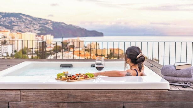 A woman relaxing in a hot tub with a glass of wine and a charcuterie board, overlooking a coastal cityscape. - Ajuda I - Penthouse by Pausa Holiday Rentals (Funchal)