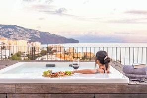 A woman relaxing in a hot tub with a glass of wine and a charcuterie board, overlooking a coastal cityscape. - Ajuda I - Penthouse by Pausa Holiday Rentals (Funchal)