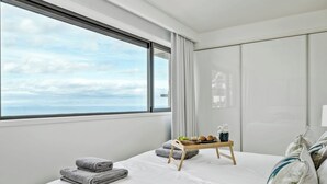 A hotel room with a large window, a bed with white linens, a bedside table with a tray of food, and a view of the sea. - Ajuda I - Penthouse by Pausa Holiday Rentals (Funchal)