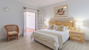 Un dormitorio con una cama, mesitas de noche, una silla de mimbre y una puerta que da a un espacio exterior.