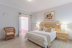 A bedroom with a bed, nightstands, a wicker chair, and a door leading to an outdoor area. - Alegria Apartment, by Pausa Holiday Rentals (Estreito da Calheta)