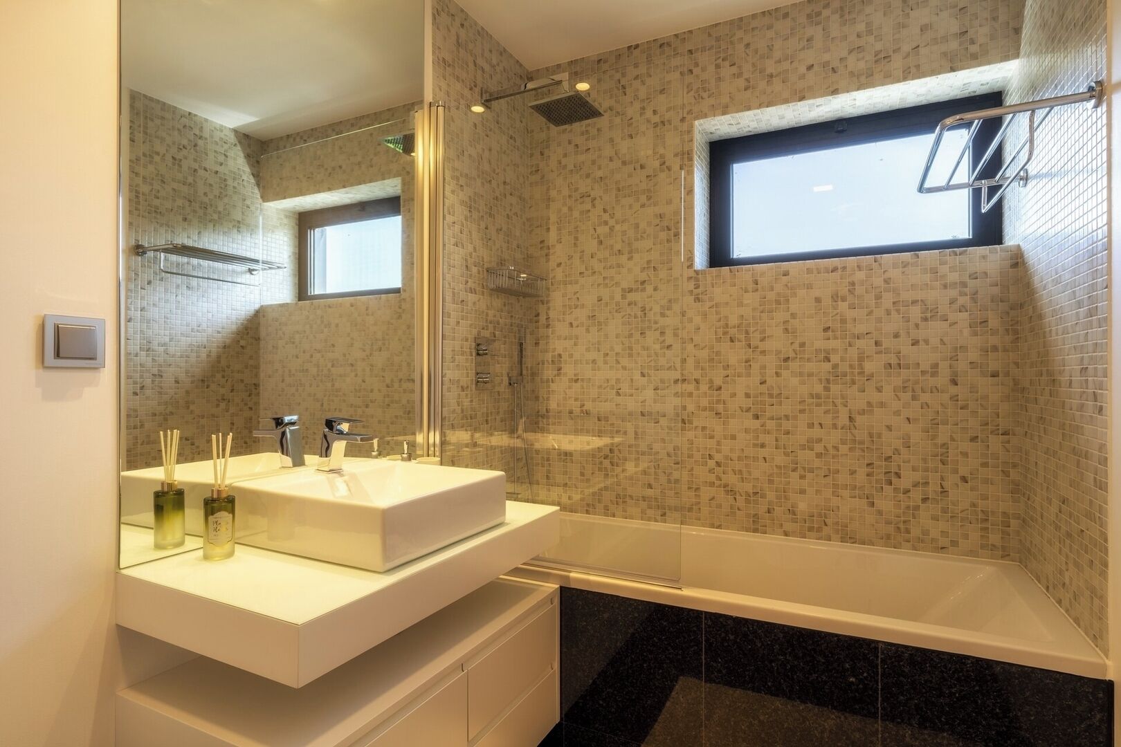 Un baño moderno con un espejo grande, un lavabo blanco, una ducha y un ventanal pequeño.