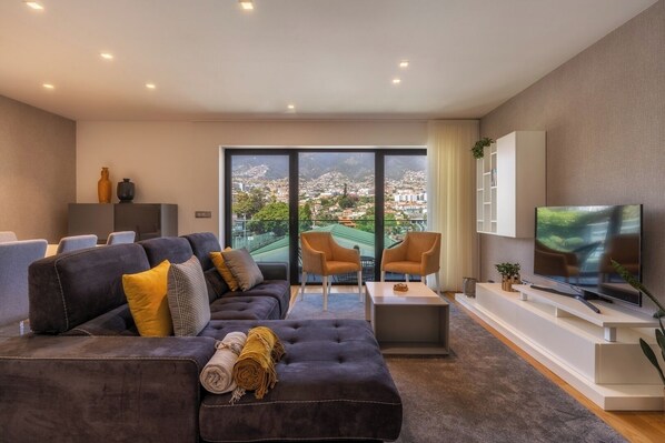 A modern living room with a large sofa, two armchairs, a coffee table, and a TV. The room has a large window with a city view. - Renala II - Apart, With Pool, by Pausa H. Rentals (Funchal)
