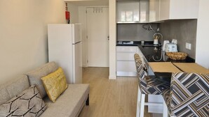 Eine kompakte Küchenzeile mit Kühlschrank, Herd und Spüle. Ein Sofa mit gemusterten Kissen und ein Esstisch mit Stühlen sind ebenfalls vorhanden.