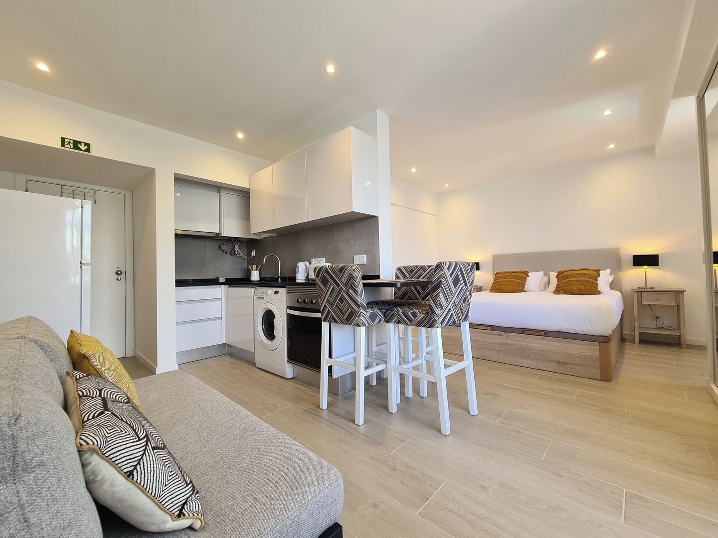 Camera d'albergo compatta con angolo cottura, zona pranzo con due sedie, letto con comodino e divano con cuscini decorati.