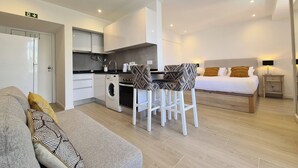 Ein kompaktes Hotelzimmer mit Küchenzeile, einem Essbereich mit zwei Stühlen, einem Bett mit Nachttisch und einer Couch mit gemusterten Kissen.