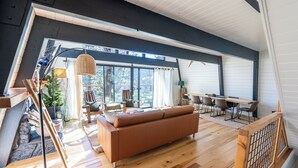 Een ruime loft-achtige woonruimte met een groot raam, een bruine bank, een eettafel en een trap.