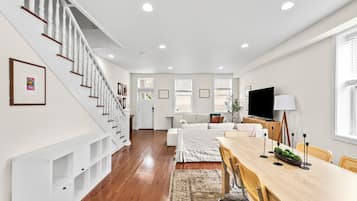 A spacious living area with a wooden staircase, a dining table with chairs, a sofa, and a television.