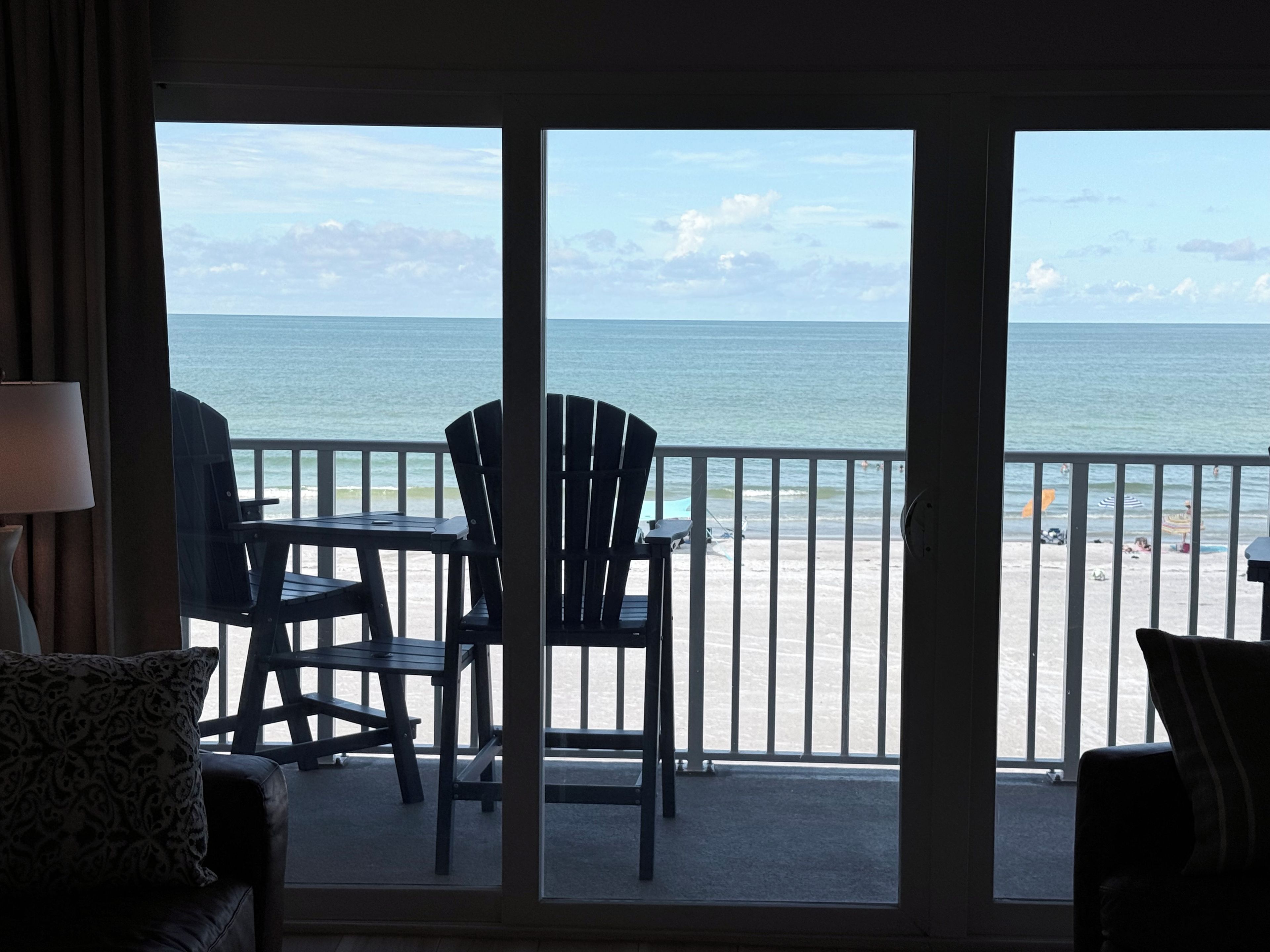 A room with a view of the beach, featuring a table and chairs, a lamp, and a window with a view of the ocean.