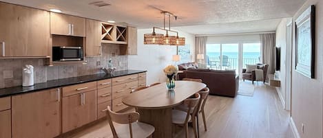 A modern kitchen with wooden cabinets and a central island, a dining area with chairs, and a living room with a sofa and a view of the ocean.