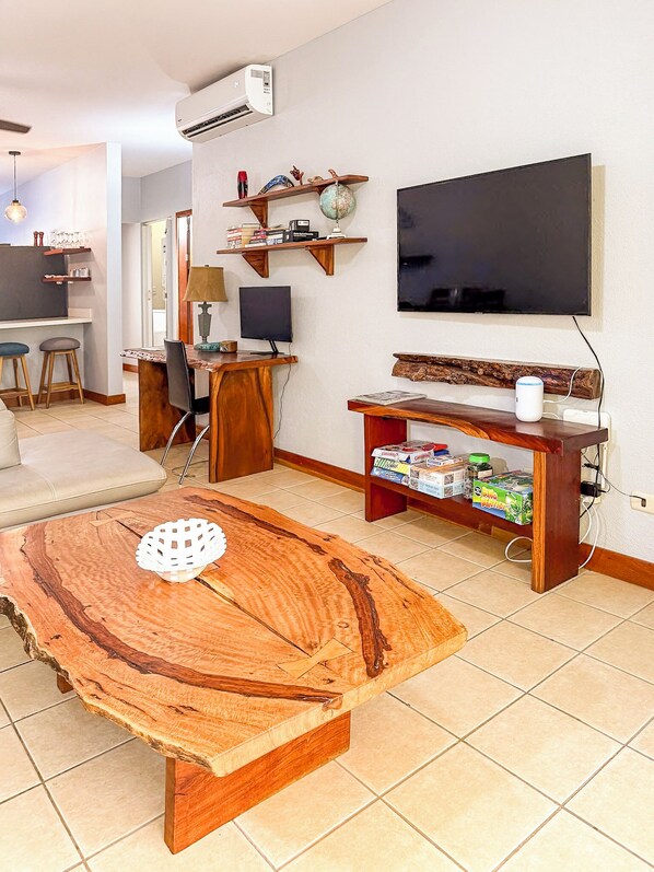 A living room with a wooden coffee table, a TV, a shelf with decorative items, and a kitchen area in the background.