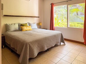 A bedroom with a bed, pillows, a nightstand, a window with curtains, and a view of greenery outside.