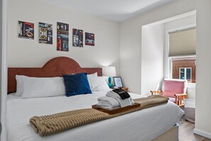 Ein Hotelzimmer mit einem großen Bett, einem Kopfteil, einem Fenster mit Jalousien, einem Sessel und Wandbildern.