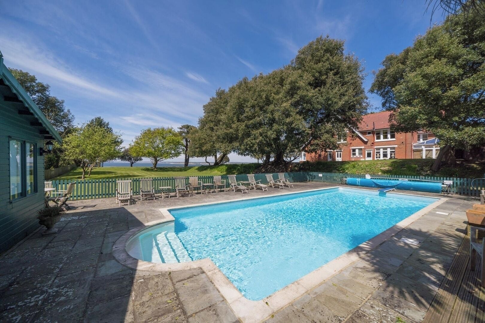 A rectangular swimming pool with a wooden deck and lounge chairs, surrounded by a wooden fence and trees, with a view of a building in the background.