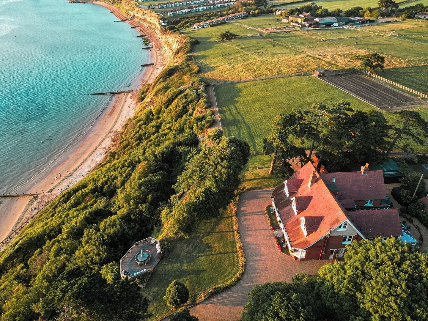 An aerial view of a coastal property with a beach, green fields, and a house with a red roof.