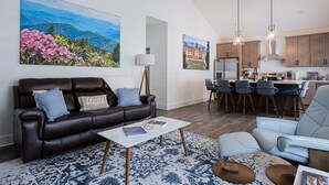 Sala de estar com sofá de couro marrom, mesa de centro branca e tapete azul estampado. Há também uma área de cozinha com armários de madeira e um balcão com banquetas.
