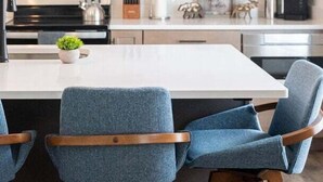 Cozinha com armários de madeira, bancada branca, coifa de aço inoxidável e um banqueta com assento azul estofado.