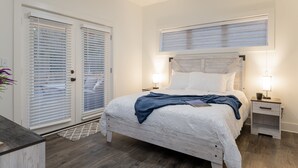 Quarto com cama, mesas de cabeceira, um criado-mudo, janela com persianas e porta com puxador.