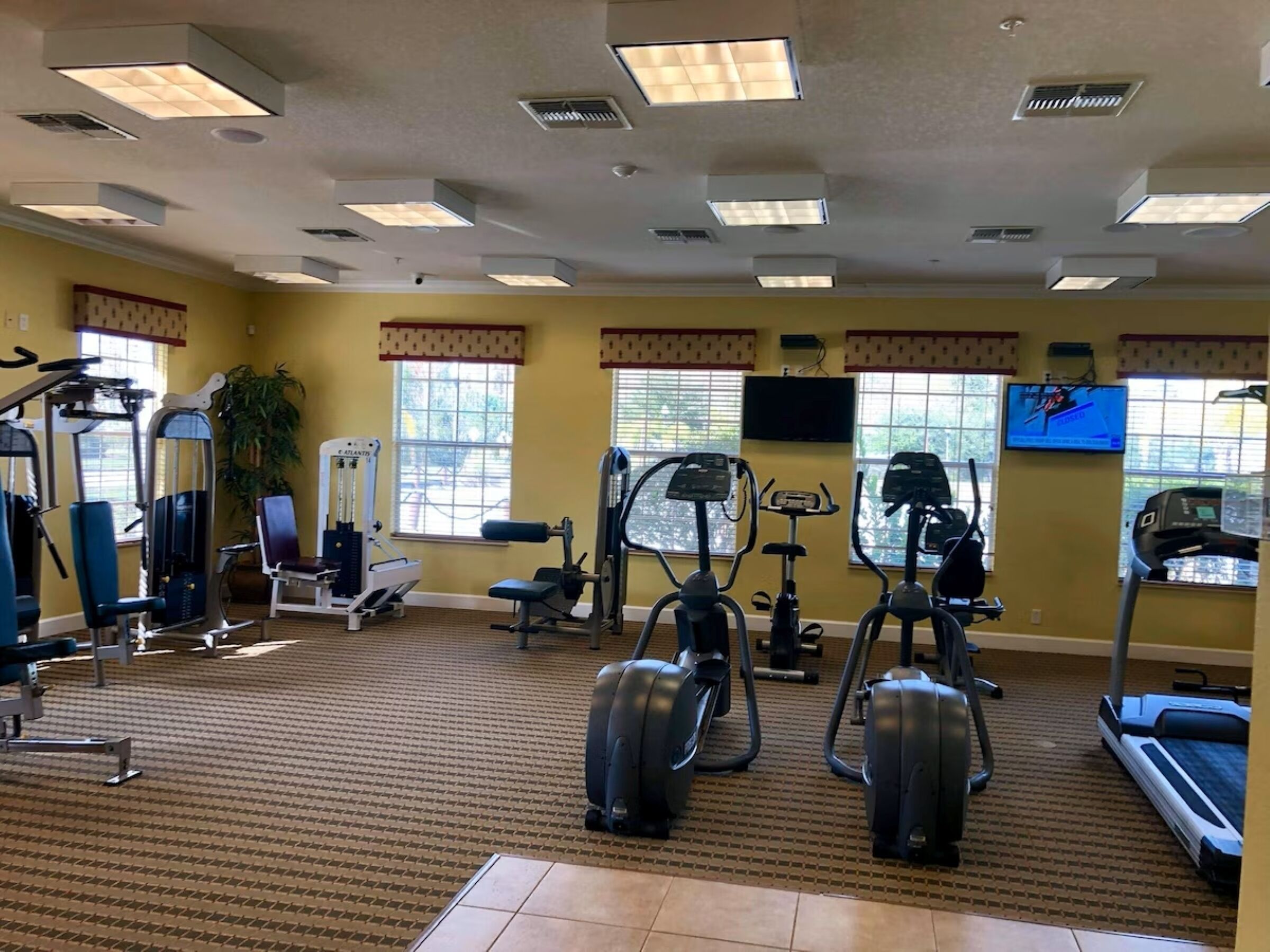Une salle de sport équipée de divers appareils d’exercice, dotée de grandes fenêtres et d’un écran de télévision.