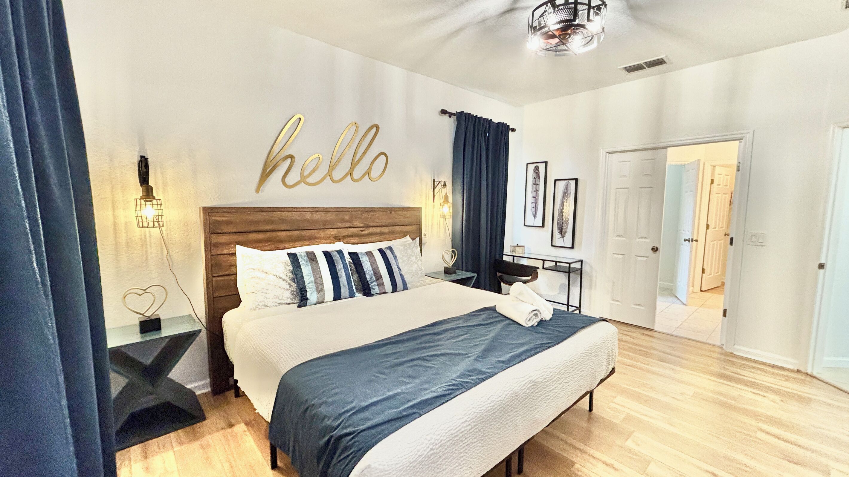 A bedroom with a wooden headboard, a bed with a blue and white duvet, a bedside table with a lamp, and a wall with a 'hello' sign.