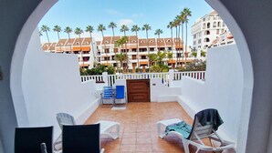 Een balkon met witte stoelen, een tafel met een fles en fruit, en uitzicht op een gebouw met palmbomen.
