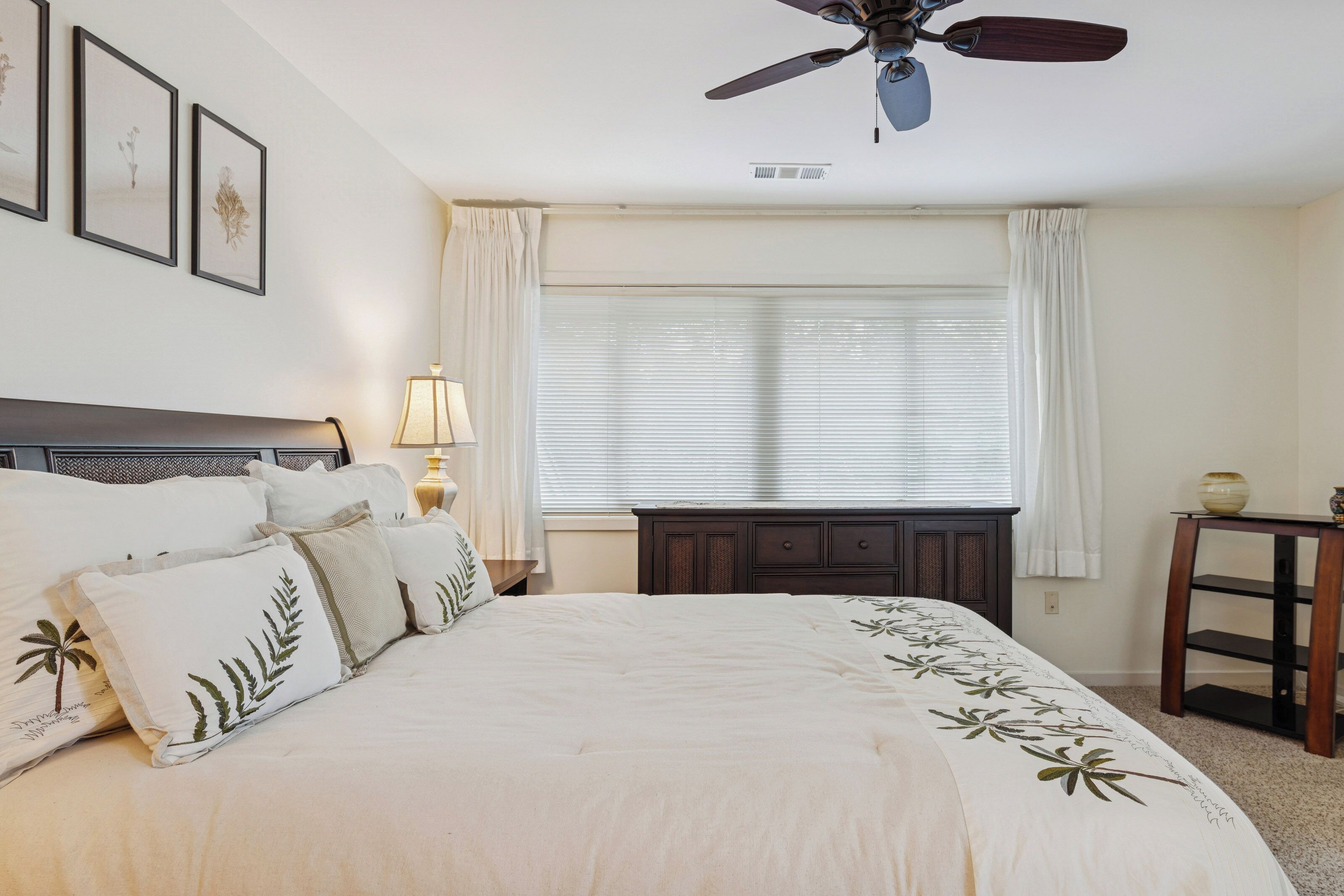 A bedroom with a bed, a ceiling fan, a window with curtains, a bedside table, and a lamp.