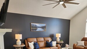 Un salon avec un canapé en cuir marron, deux tables d’appoint avec des lampes, un ventilateur de plafond, un tableau représentant un paysage encadré et un mur de couleur sombre.