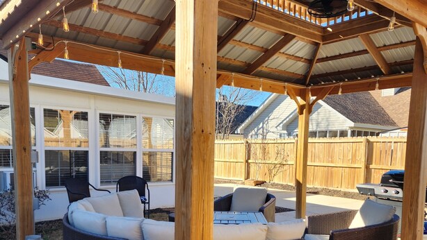 Un espace extérieur aménagé avec un coin salon abrité, comprenant un grand canapé de couleur foncée et des fauteuils plus petits, une bouteille de propane et un barbecue à l’arrière-plan.