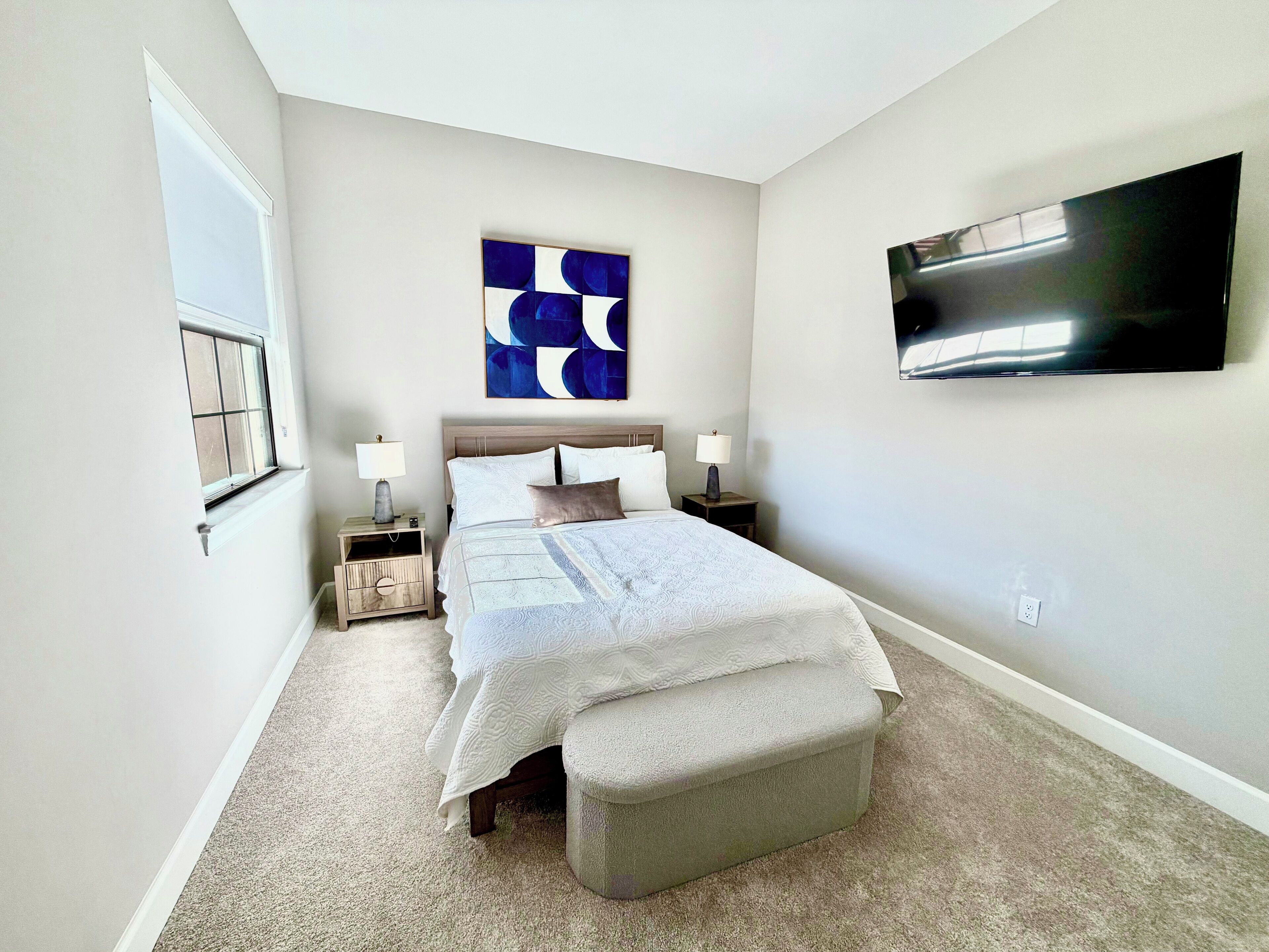 A bedroom with a bed, two bedside tables, a bench, a wall-mounted TV, and a window.